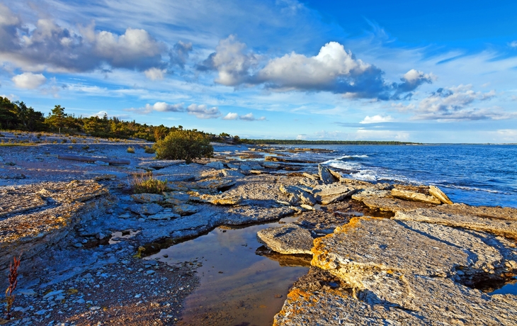 Steinige Küstenlandschaft mit Wolken am blauen Himmel und Meer im Hintergrund.