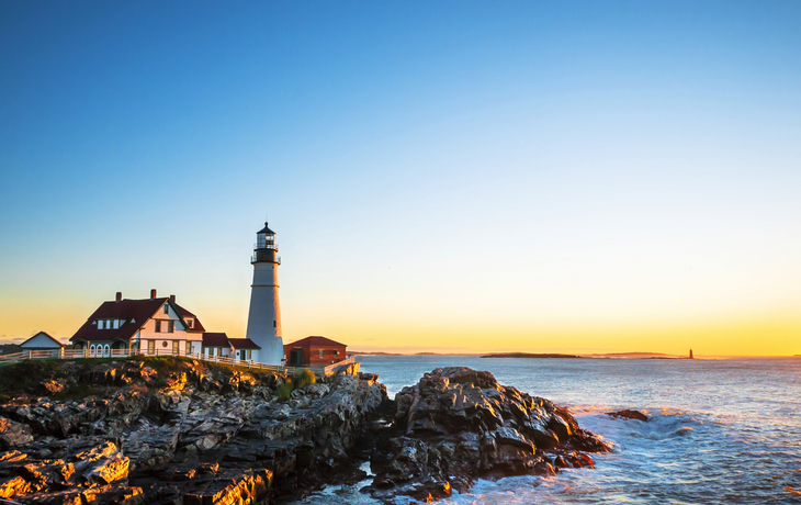 Leuchtturm auf Felsenküste bei Sonnenuntergang, ruhige See im Hintergrund.