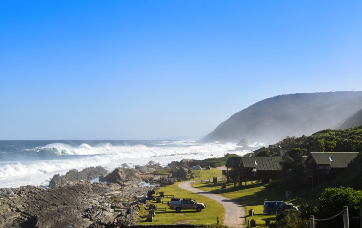 Küstenlandschaft mit Wellen, Straße und Hütten am Meer bei klarem Himmel.