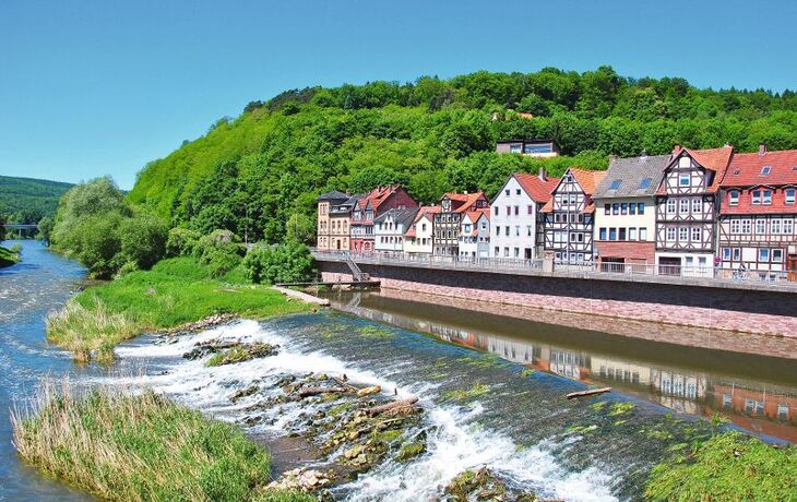 Fachwerkhäuser am Flussufer mit grüner bewaldeter Hügellandschaft im Hintergrund.