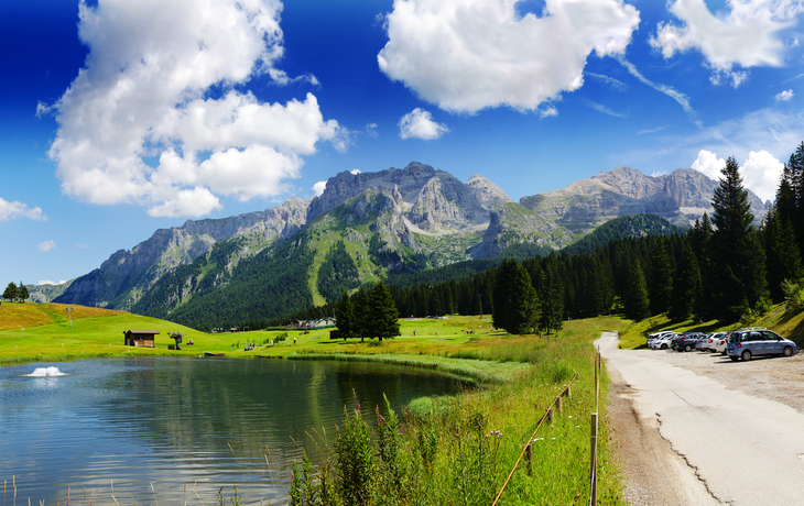 Berglandschaft mit See, Wiese, Bäumen und geparkten Autos unter bewölktem Himmel.
