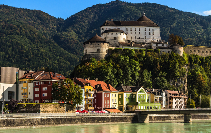 Festung auf einem Hügel mit bunten Häusern am Flussufer im Vordergrund.
