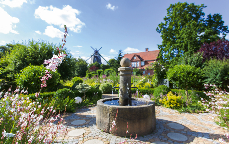 Brunnen im Park mit Mühle und blühenden Blumen im Hintergrund.