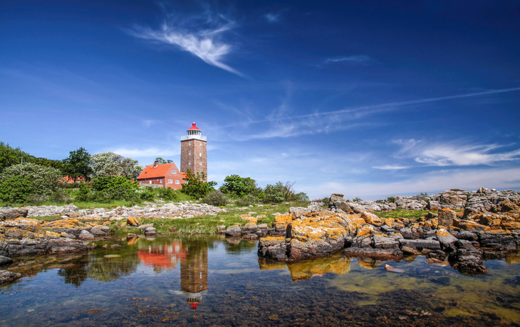 Ein Leuchtturm mit roten Dachgebäuden neben Felsen und Wasser, blauer Himmel darüber