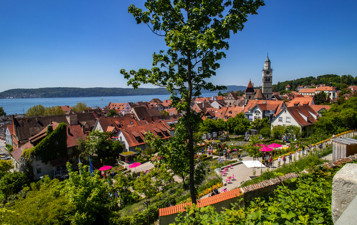 Stadtansicht mit Kirche und See im Hintergrund, Bäume im Vordergrund, sonniger Tag.