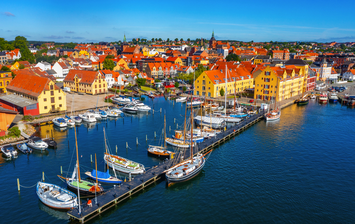 Hafenstadt mit Segelbooten und bunten Häusern am Wasser unter blauem Himmel.