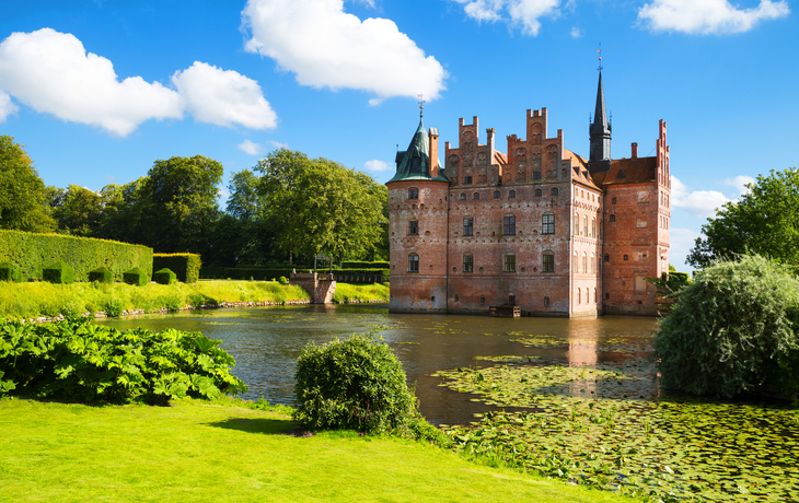 Backsteinschloss mit Wassergraben und Garten bei klarem Himmel.