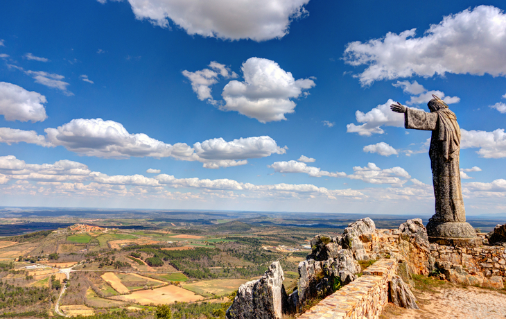 Statue auf Klippe mit weitem Blick über ländliche Landschaft und Wolkenhimmel.