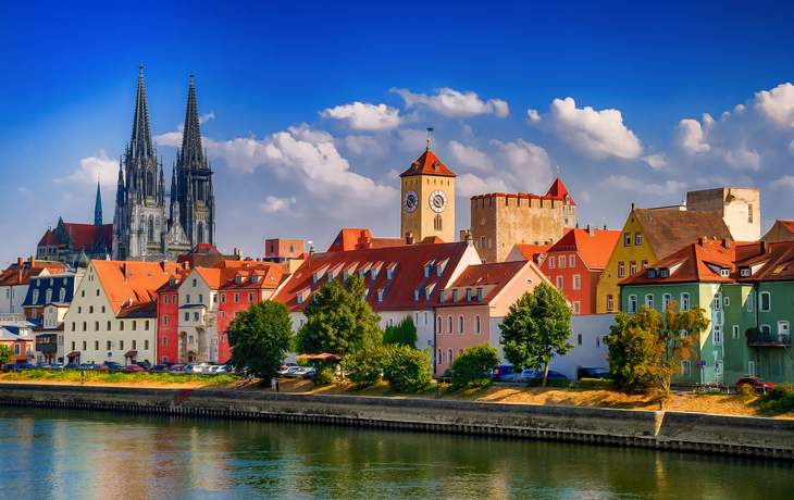 Bunte Häuserzeile am Fluss mit Kathedrale und blauem Himmel im Hintergrund.
