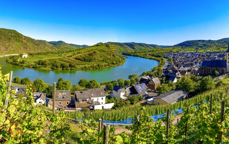 Blick auf malerisches Flusstal mit Dorf und Kirche, umgeben von Hügeln und Weinreben.