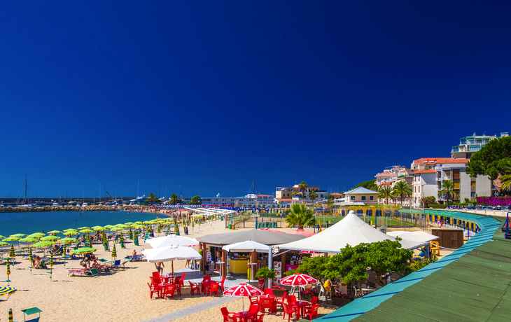 Beliebter Sandstrand mit Sonnenschirmen und Palmen, blauer Himmel im Hintergrund.